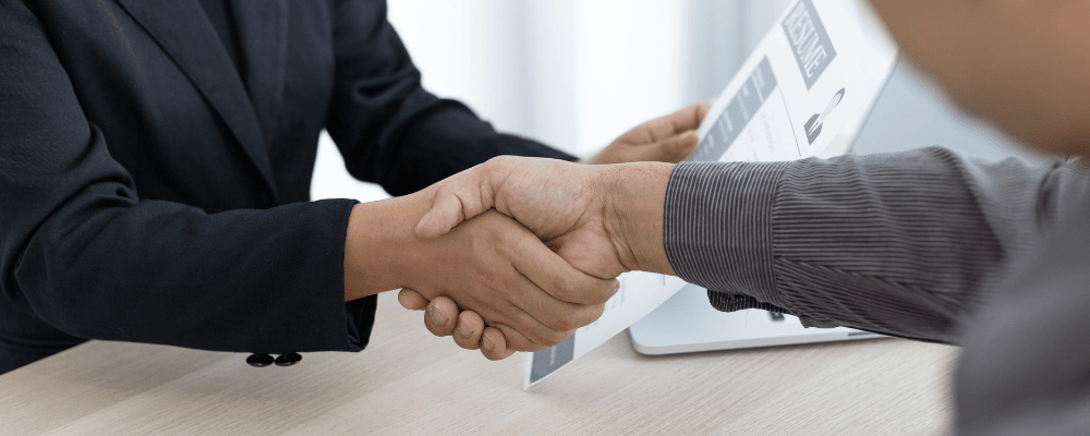 Zwei Personen reichen sich die Hand über einem Tisch, im Hintergrund ein Lebenslauf – Symbol für Bewerbungsgespräch oder Jobzusage.