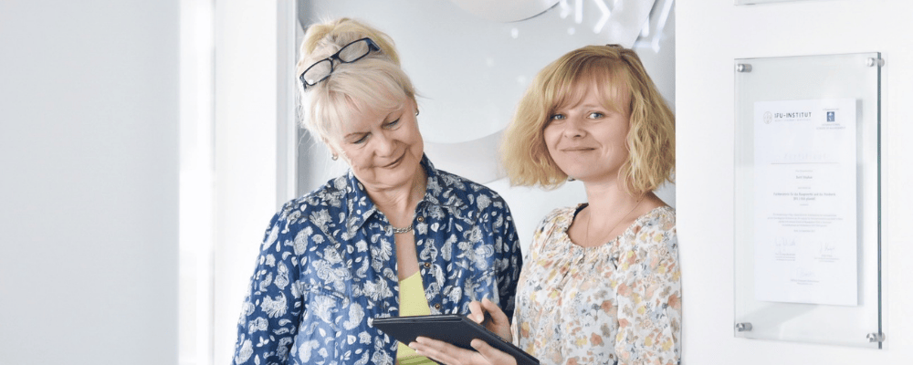 Zwei Mitarbeiterinnen im Gespräch vor einer Urkunde an der Wand. Eine zeigt der anderen etwas auf einem Tablet. Beide lächeln.